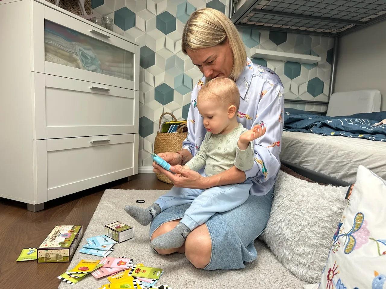 Maminka s malým dítětem sedí na zemi v dětském pokoji a společně si prohlížejí mluvící karty Kinderspeech pro hravé učení.