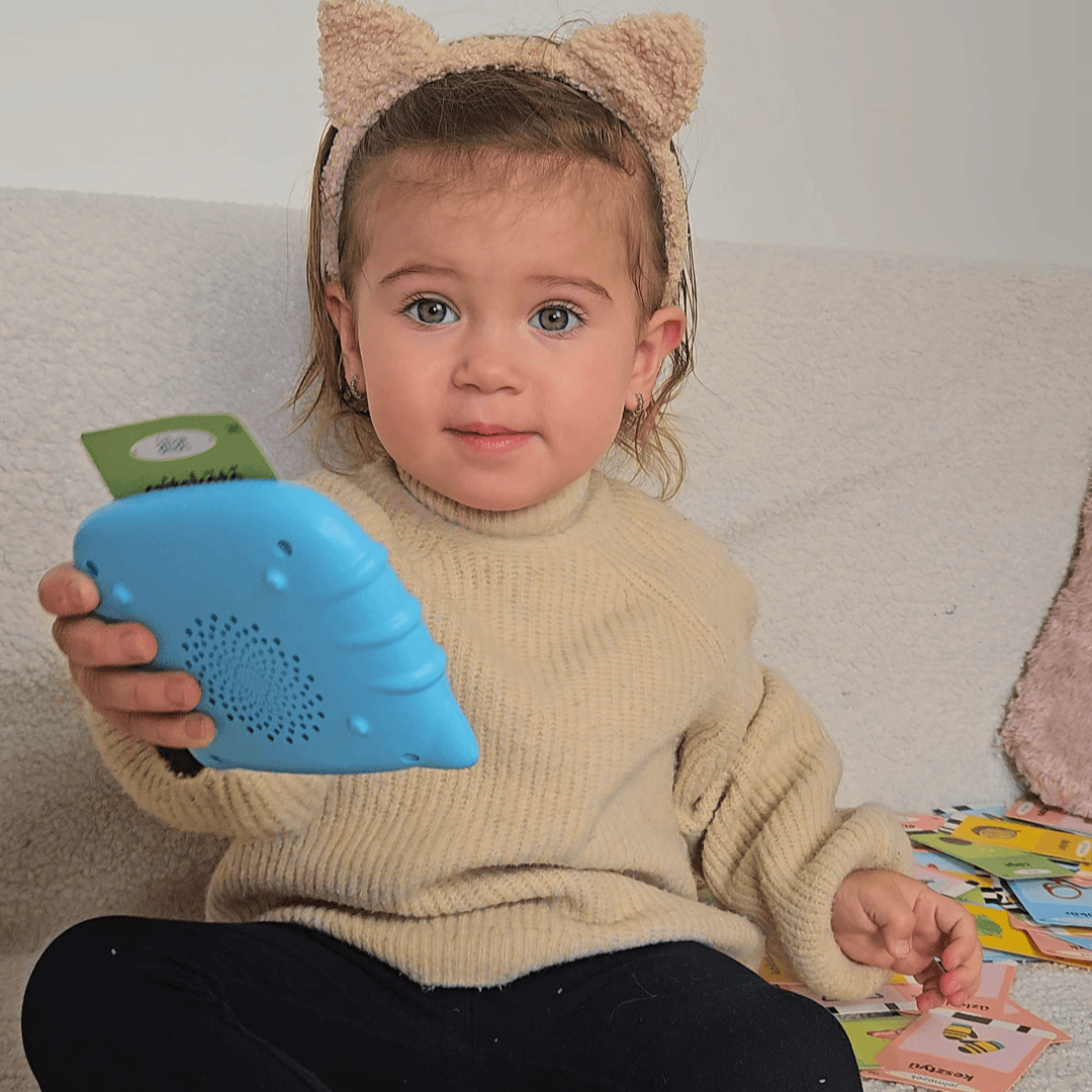 Malá holčička s kočičími oušky si hraje s modrou mluvící čtečkou Kinderspeech pro zábavné učení nových slovíček.